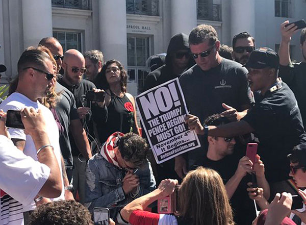 Milo Yiannopoulos de rodillas con los manifestantes que lo rodean.